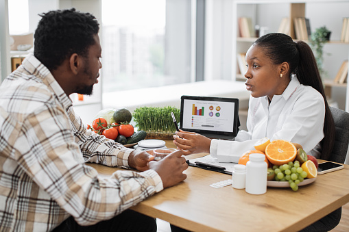 Experienced professional displaying nutrition data graphically via tablet for multicultural man in doctor's workplace. Female expert in diet motivating client to right food choices during treatment.