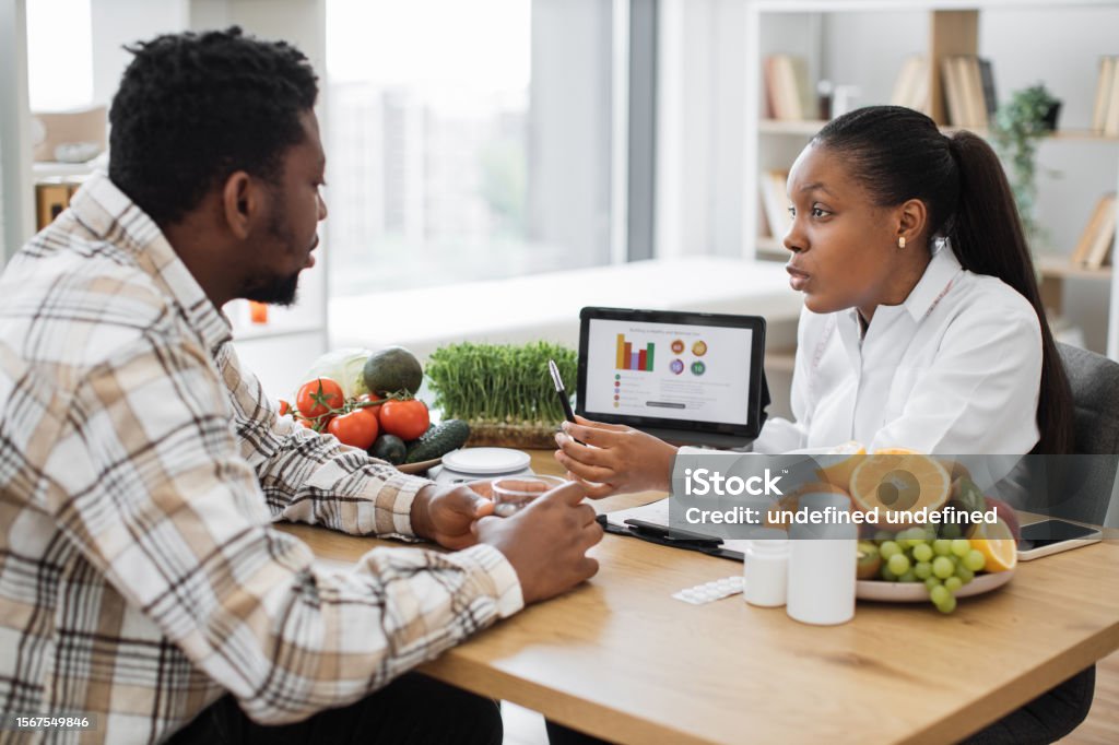 Expert in diet displaying client's data on tablet via graphs Experienced professional displaying nutrition data graphically via tablet for multicultural man in doctor's workplace. Female expert in diet motivating client to right food choices during treatment. Nutritionist Stock Photo