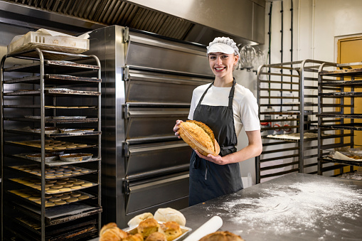 A young woman is standing in front of an industrial oven, she is wearing a white cap, a white t-shirt and a dark apron. She is smiling at the camera and holding a freshly baked loaf in her hands.