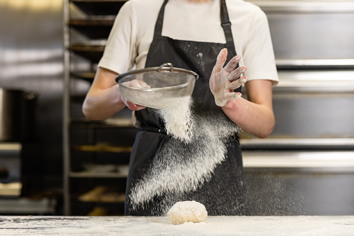 A baker is in an industrial kitchen, she is sprinkling flour through a sieve onto a ball of dough which is on a flour covered stainless steel table. In the background are cooling racks and ovens.