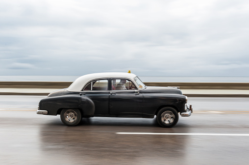 HAVANA, CUBA - OCTOBER 21, 2017: Old Car in Havana, Cuba. Retro Vehicle Usually Using As A Taxi For Local People and Tourist. Caribbean Sea in Background. Panning Black Color Car