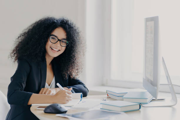 istock Cheerful student, glad expression, notes in notebook, wears transparent glasses, black formal wear, at desk with books and computer. 1567613625