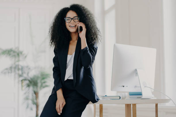 istock Satisfied dark-skinned businesswoman on break, leaning on white table with computer in her office, talking on her cellphone, wearing glasses and an elegant suit, consulting with a partner. 1567613630