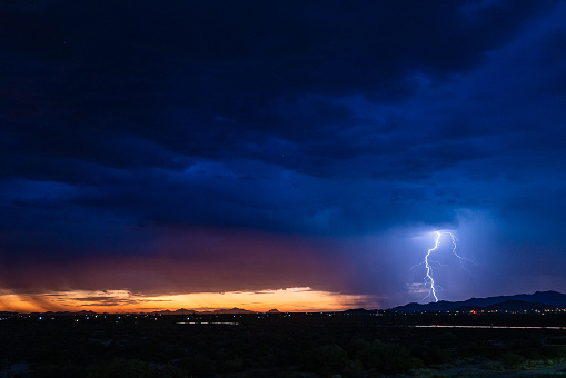 An intense monsoon storm is colored by the setting sun to the west in the Sonoran Desert landscape. Lightning strikes to the right, while rain can be seen falling on the left.