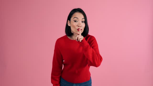 Happy Asian woman in red sweater showing silence gesture at camera