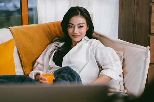 Young Asian woman lying on the sofa bed enjoying the weekend, watching movie on TV and eating a food at home in the evening, relaxation time indoor a house, Young woman watching news on television