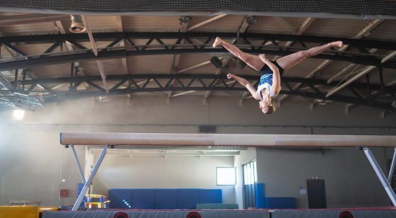 Female gymnast doing a complicated trick on balance beam in gym. Sport and endurance concept.