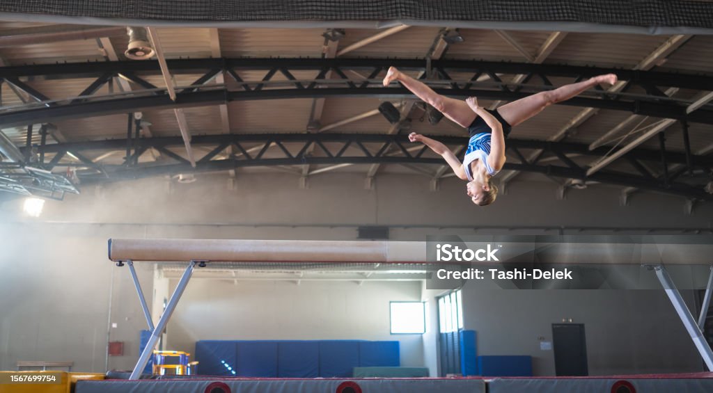 Female Gymnast On Balance Beam Female gymnast doing a complicated trick on balance beam in gym. Sport and endurance concept. Accuracy Stock Photo