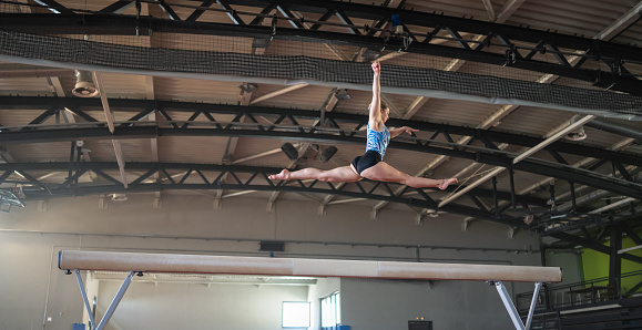 Female gymnast doing a complicated trick on balance beam in gym. Sport and endurance concept.