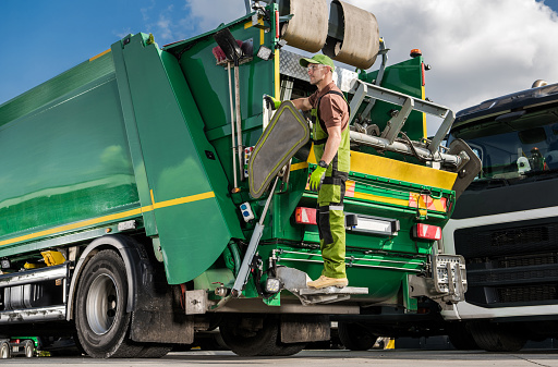 Caucasian Modern Garbage Truck Worker Riding on a Rear Side of the Vehicle. Waste Management Industry Job. Sorting and Recycling Theme.