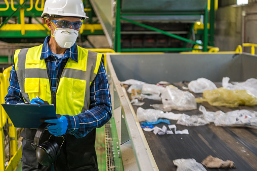 Waste Management Sorting Facility Caucasian Worker Preparing Documentation. Wearing Air Pollution Mask, Eyes Safety Glasses and a Hard Hat.