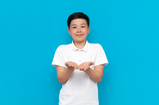 little asian boy in white t-shirt holds his hands in front of him and begs on blue background, korean child holds empty palms on isolated background