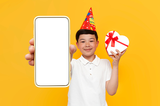 little asian boy in party hat holds heart-shaped gift and shows blank smartphone screen on yellow background, korean child wishes happy birthday online on the phone