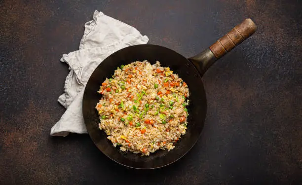 Photo of Authentic Chinese and Asian fried rice with egg and vegetables in wok top view, rustic concrete table background. Traditional dish of China