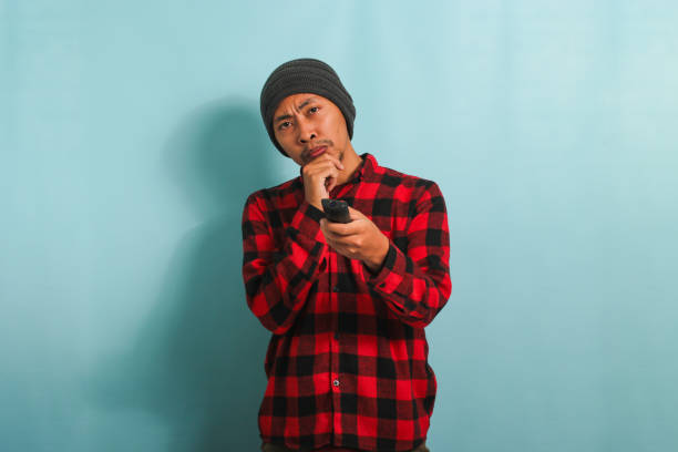istock Bored young Asian man holding a remote control while watching TV, isolated on a blue background 1567926854