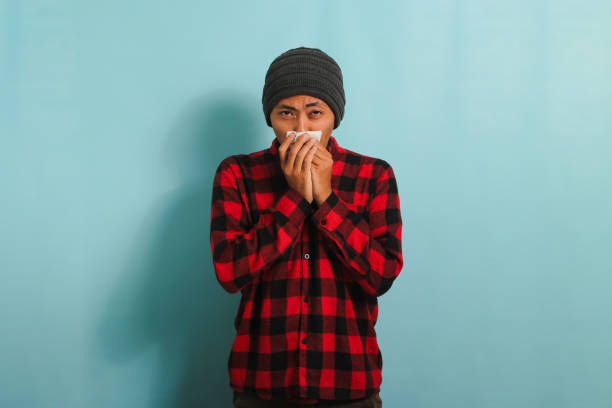 istock Young Asian man is sneezing into tissue, suffering from cough, and flu, isolated on blue background 1567933369