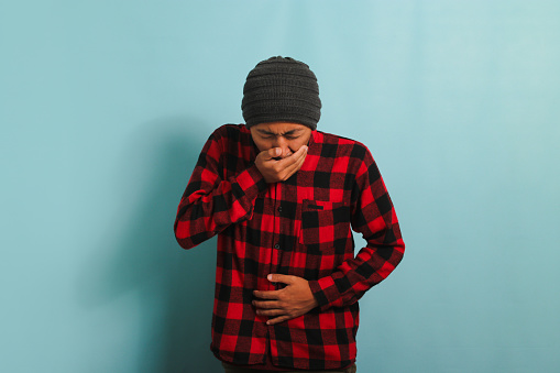 An unhealthy young Asian man with a beanie hat and a red plaid flannel shirt is moaning suffering from a stomach ache, bending over and holding his hands on his belly, isolated on blue background