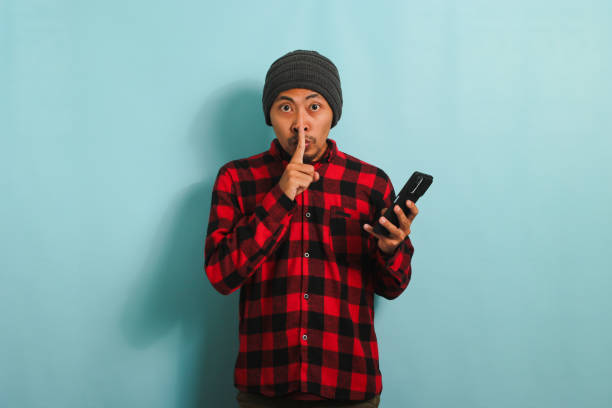 istock Young Asian man makes silent gesture to keep quiet while holding phone, isolated on blue background 1567936542