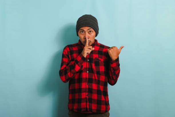 istock Young Asian man is making silent gesture, while pointing at copy space, isolated on blue background 1567937050