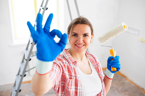 Beautiful woman holding roller painter doing ok sign with fingers while doing repair in room.