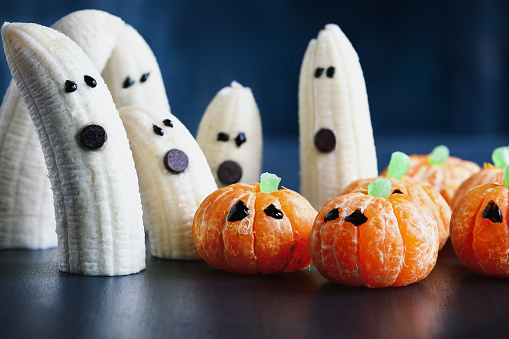 Halloween cute pumpkin orange fruit and scary banana ghosts monsters with chocolate faces. Healthy dessert snack with funny faces for child's party decoration. Selective focus on center food with blurred background.