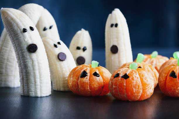 Halloween cute pumpkin orange fruit and scary banana ghosts Halloween cute pumpkin orange fruit and scary banana ghosts monsters with chocolate faces. Healthy dessert snack with funny faces for child's party decoration. Selective focus on center food with blurred background. eye candy stock pictures, royalty-free photos & images