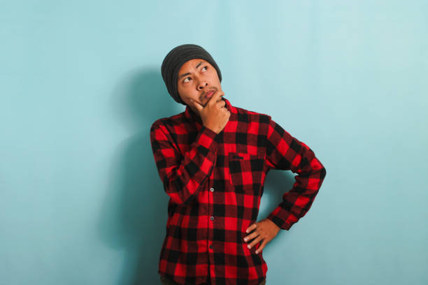 istock Pensive young Asian man keeps his hand on his chin, looking at copy space, isolated on blue backdrop 1567984192
