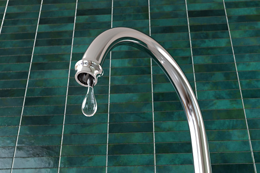 Silver kitchen water tap (faucet) dripping a large drop of water in front of dark green tiles wall. Illustration of the concept of water saving and pipe leakage