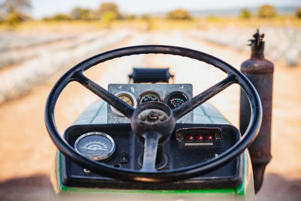 A close-up view of a tractor steering wheel with various gauges and dials mounted on the seat in a rural outdoor field setting