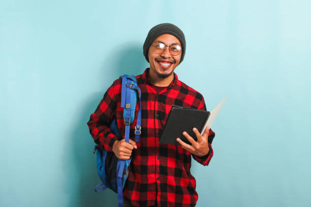 istock Young Asian student man wearing a backpack is holding books while standing against a blue background 1567994486