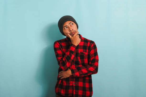 istock Pensive young Asian man keeps his hand on his chin, looking at copy space, isolated on blue backdrop 1568001123