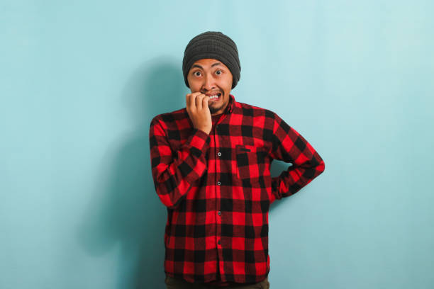 istock Young Asian man bites his fingernails, looking nervously at the camera, isolated on blue background 1568001127