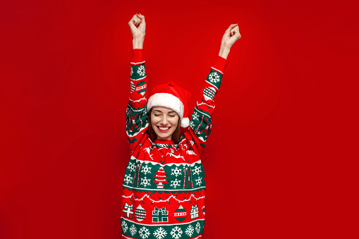 young cute girl in christmas sweater and santa hat rejoice in victory and shout on red background, winner woman in new year clothes celebrates success and good luck with raised hands