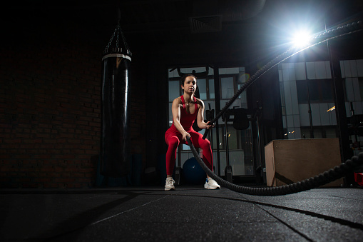 young sports girl in red sportswear trains with ropes in a black dark gym, motivated woman on a fitness training goes in for sports, copy space