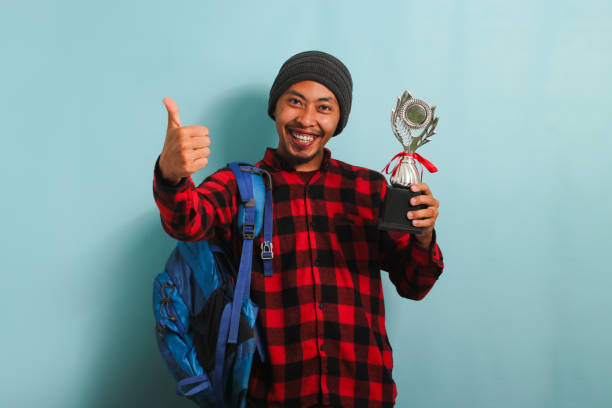 istock Excited Young Asian man student giving thumbs-up while holding a trophy, isolated on blue background 1568007631