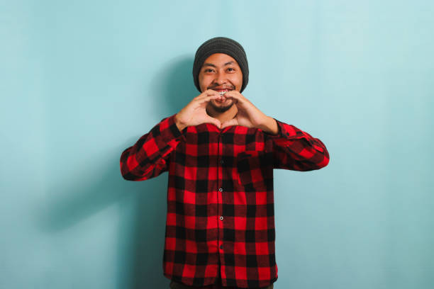 Smiling young Asian man shows a love gesture with his hand while standing against a blue background
