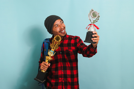 Happy young Asian man student wearing a backpack, beanie hat, and red plaid flannel shirt, lifting up his trophies, rejoicing in his success and achievement, isolated on a blue background.