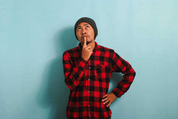 istock Pensive young Asian man keeps his hand on his chin, looking at copy space, isolated on blue backdrop 1568041137