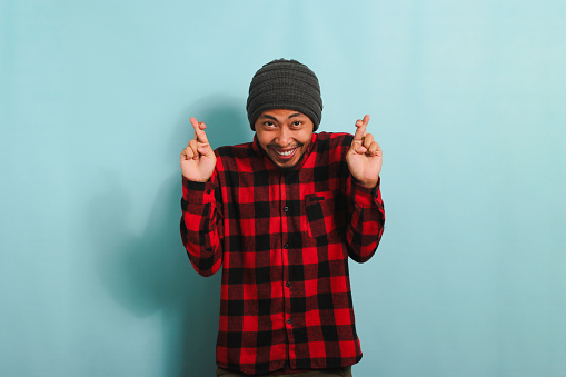 An excited young Asian man with a beanie hat and a red plaid flannel shirt crosses his fingers and rejoices, hopeful that his dreams have come true while standing against a blue background