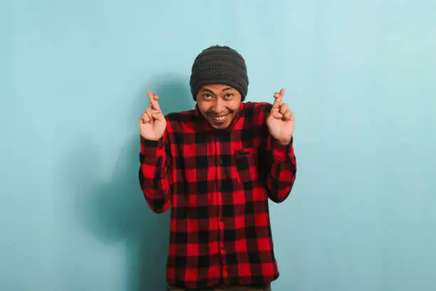 Photo of Excited young Asian man makes finger crossed gesture, isolated on blue background