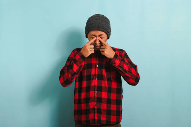 Young Asian man rubbing his swollen eyes due to dust allergy and itching isolated on blue background Young Asian man with beanie hat and red plaid flannel shirt experiencing discomfort with contact lenses, rubbing his swollen eyes due to dust allergy and itching, isolated on a blue background human eye scratching allergy rubbing stock pictures, royalty-free photos & images