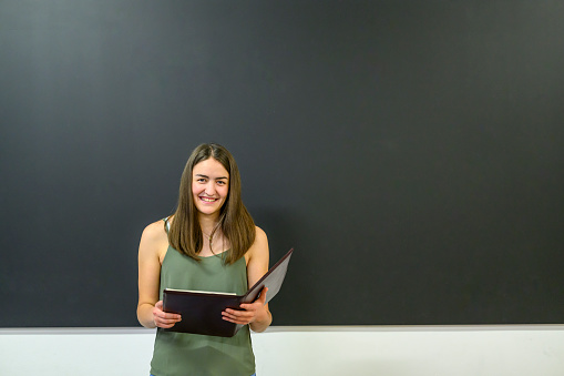Happy smiling female instructor standing with a thin file at the blackboard in a classroom