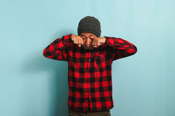 Young Asian man rubbing his swollen eyes due to dust allergy and itching isolated on blue background Young Asian man with beanie hat and red plaid flannel shirt experiencing discomfort with contact lenses, rubbing his swollen eyes due to dust allergy and itching, isolated on a blue background human eye scratching allergy rubbing stock pictures, royalty-free photos & images