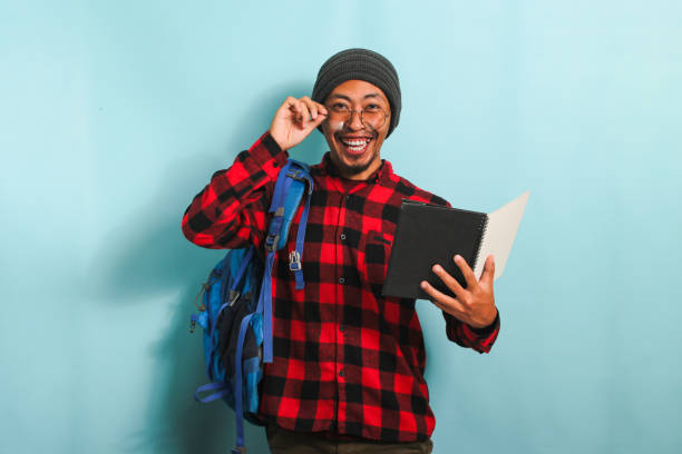 istock Young Asian student man wearing a backpack is holding books while standing against a blue background 1568050281