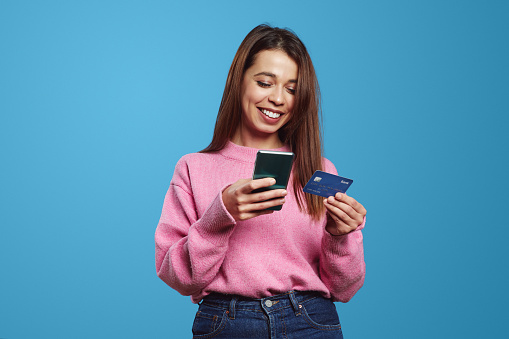 Excited young girl paying online, holding smartphone and credit card, enter info in application on mobile phone, standing against blue background