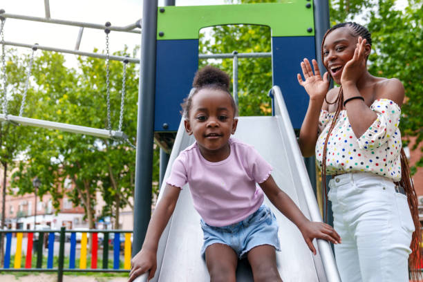Joyful African Toddler: Radiant Smiles at Playground, Surprises Mother Amidst Lush Greenery.
