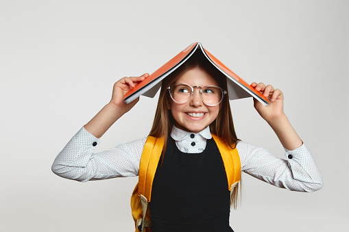 Funny clever schoolgirl wearing eyeglasses and holding open textbook on head smiling and looking away while doing homework against white background