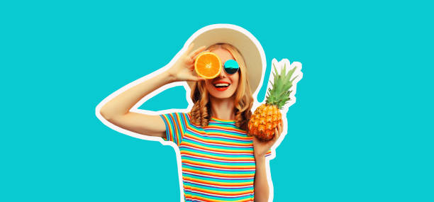 Summer portrait of happy smiling young woman with pineapple and slice of orange, fresh tropical juicy fruits, wearing straw hat, sunglasses on blue background stock photo