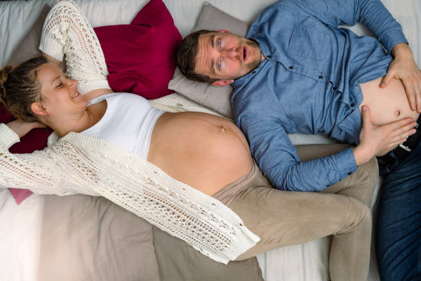 будущие родители лежат на диване, а беременный отец держится за живот. яркий кадр. - baby animal nest newborn lying down стоковые фото и изображения