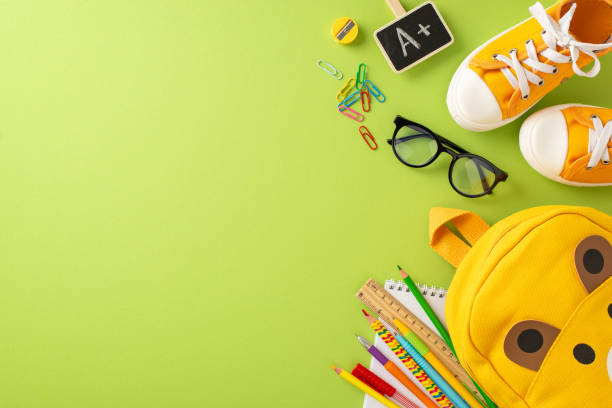 istock Get ready for elementary school. Top view of bear-shaped backpack, colored markers, sketchbook, art supplies, clips, glasses, A+ grade symbol, sneakers on soft green backdrop with space for text or ad 1568181654